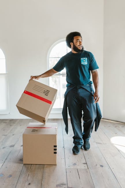 A man with a curly beard and dark curly hair, wearing a navy blue t-shirt with a blue and white logo on the chest, is inside a well-lit room with white walls and wooden flooring. He is holding a cardboard box with red tape around it, preparing to place it on the floor or a stack of similarly packed boxes in the process of home relocation. Behind him, there is a large arched window allowing natural light to fill the space. The room appears to be part of a residential property, with the ongoing moving process facilitated by Removals Swiss Cottage, as indicated by the context of the page about house moves on tricky streets. The scene captures the packing and lifting stages of furniture transport and packing during a professional moving service, with the man positioned near the window and the boxes arranged on the wooden floor.