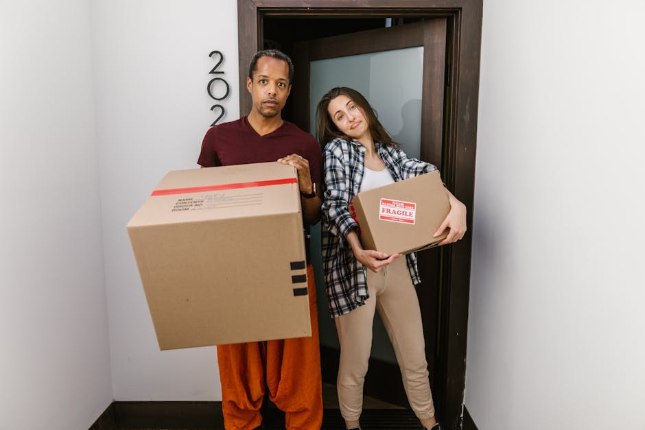 Two movers from Swiss Cottage Removals stand in a doorway of a residential building, each holding a cardboard moving box. The male mover, dressed in a maroon T-shirt and orange trousers, holds a large box with black tape on the edges, while the female mover, wearing a white top, plaid shirt, and beige trousers, carries a smaller box with a red 'Fragile' label. Behind them, the interior corridor features a plain white wall and a dark wooden door frame, with the door slightly open. The team appears to be engaged in a home relocation process, preparing to load furniture and packaging materials into a van, supported by the visual cues of packing and handling equipment nearby. The lighting is bright and even, emphasizing a professional moving operation consistent with services offered by Swiss Cottage Removals focusing on complex street access and careful furniture transport.